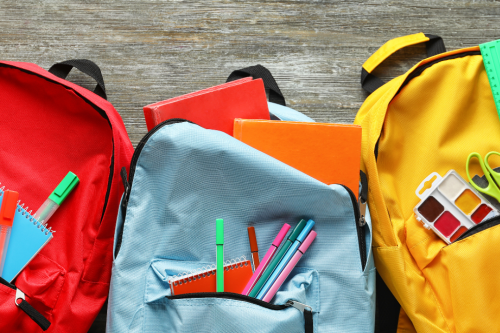 Três mochilas coloridas abertas com material escolar visível, incluindo cadernos, canetas e réguas